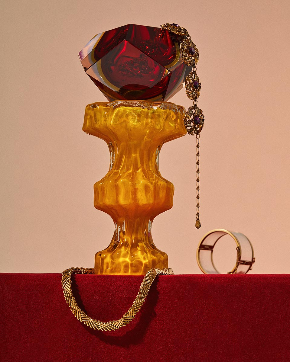 Still life with a red faceted glass container on an amber pedestal, complemented by a gold necklace and bracelet on a red surface.