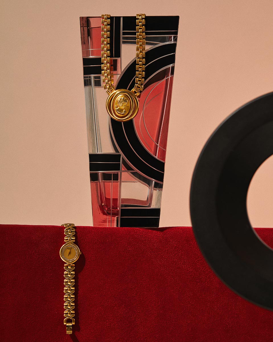 Still life shot featuring a golden wristwatch and necklace draped over a patterned glass vase, set against a red cloth.