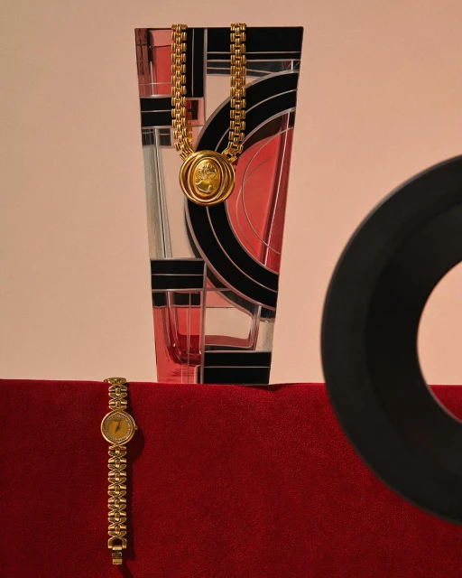 Still life shot featuring a golden wristwatch and necklace draped over a patterned glass vase, set against a red cloth.