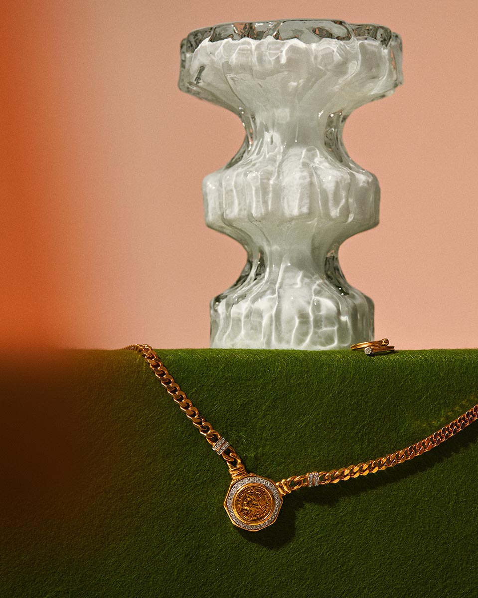 Elegant studio shot of a gold medallion necklace on a green velvet surface next to a clear glass decorative vase against a peach background.