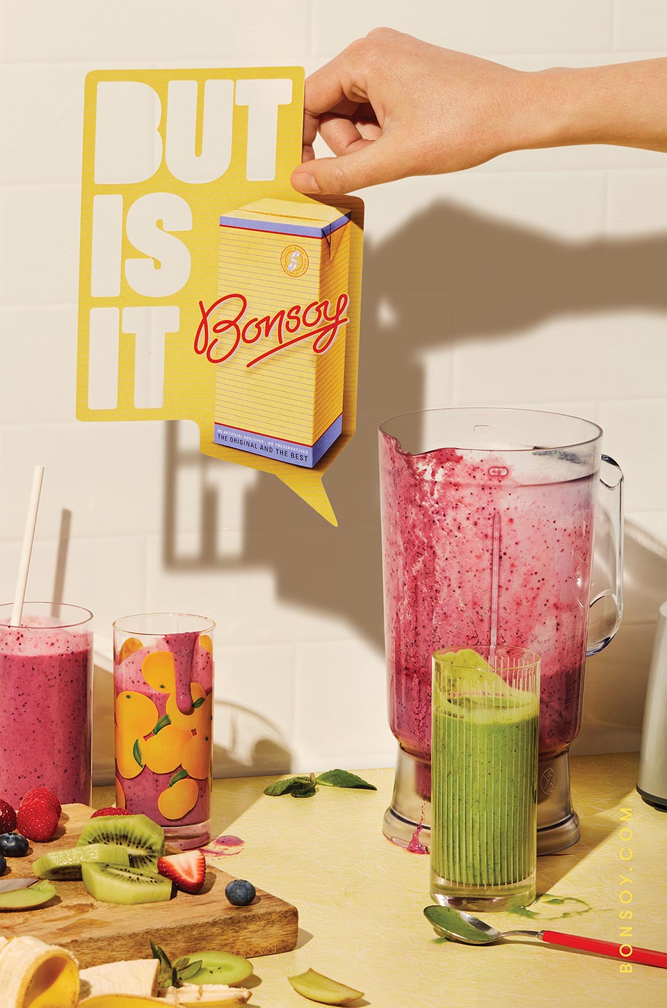 Still life of various smoothies, fruits, and a hand holding a Bonsoy advertisement sign in a studio with soft, diffused lighting.