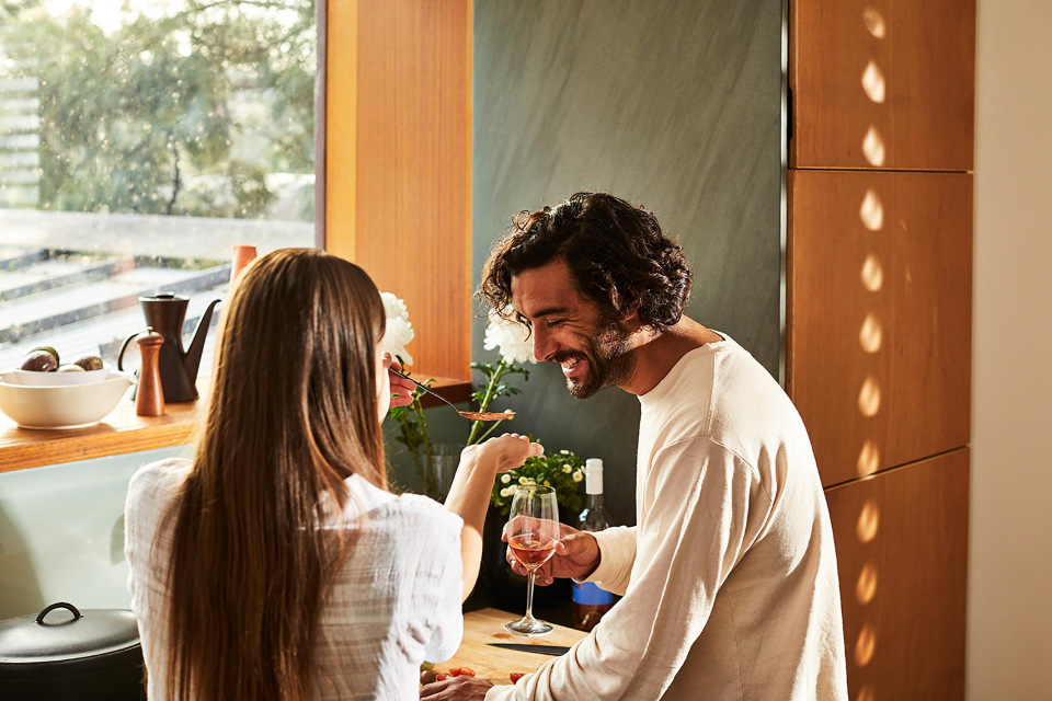 A smiling man and woman in a kitchen preparing food with a glass of wine, enjoying a sunlit interior setting.