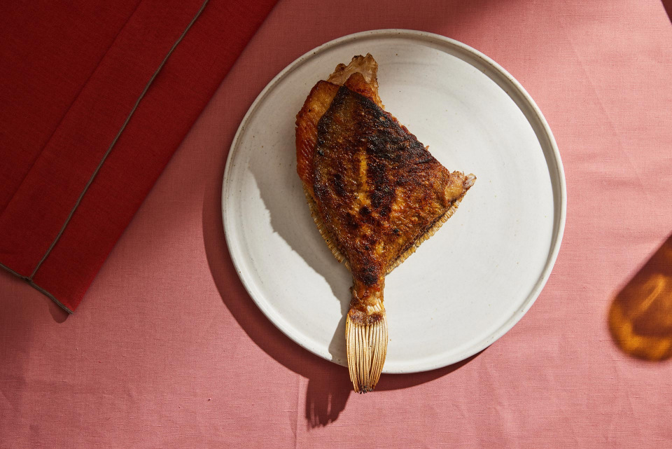 Overhead shot of a cooked flounder on a white plate. The fish sits on a pink linen tablecloth next to a red cushion.