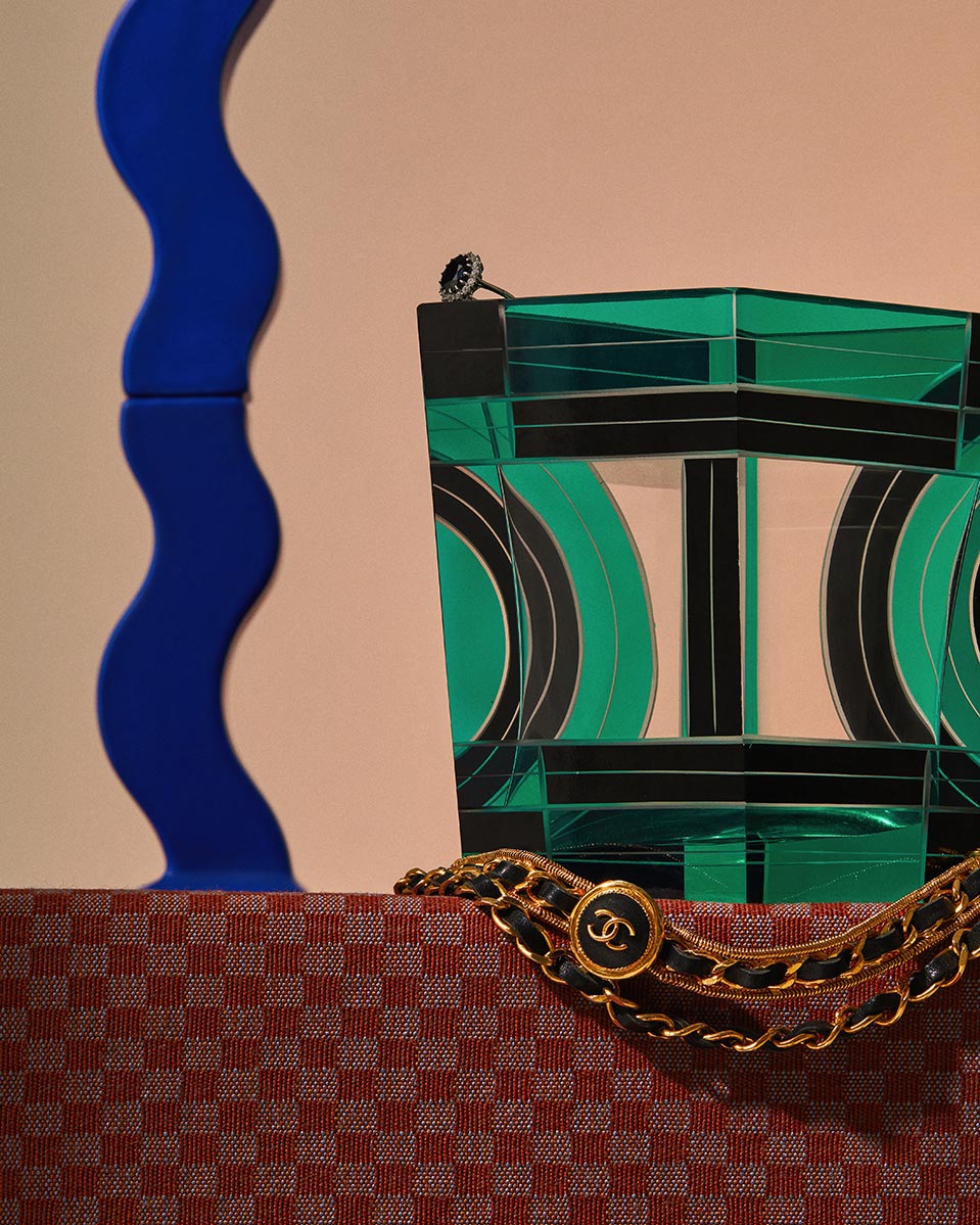 Still life photograph of a geometric glass sculpture, a ring, a gold and black belt, and a blue wave sculpture on a red checkered surface.