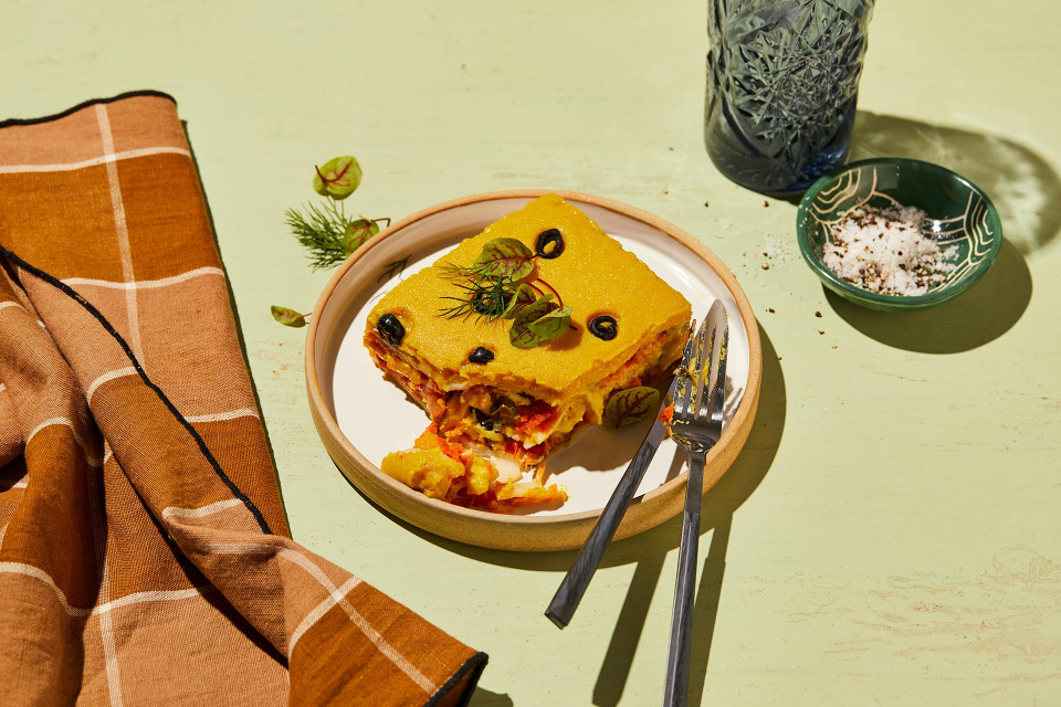 Slice of layered vegetable dish with tomato, olives, greens on a plate with forks, set on a pale green surface with a brown towel.