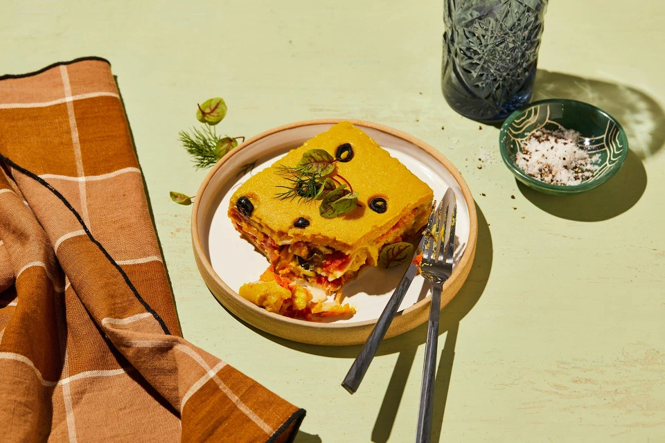 Slice of layered vegetable dish with tomato, olives, greens on a plate with forks, set on a pale green surface with a brown towel.