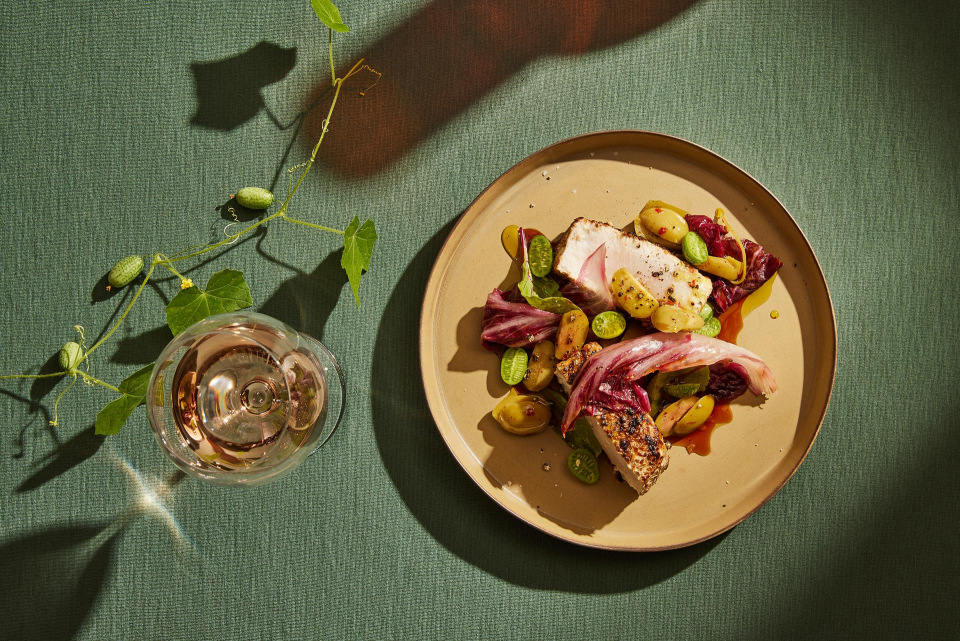 Overhead view of seared tuna steak and vegetable salad on plate with wine glass, casting shadows on green textured backdrop.