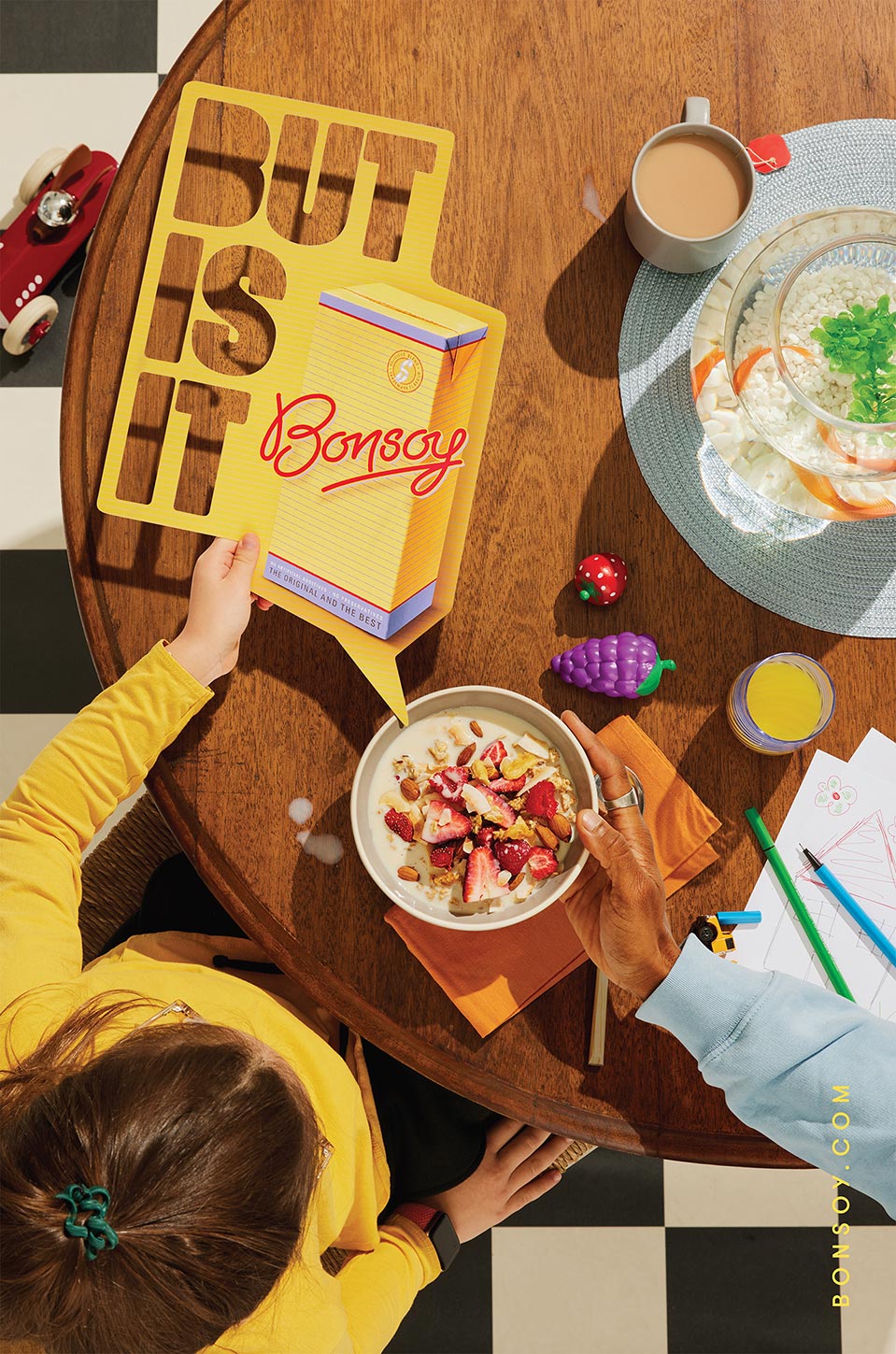 Overhead view of child and adult hands on a wooden table with cereal bowl, Bonsoy carton speech bubble, drinks, and drawing materials.