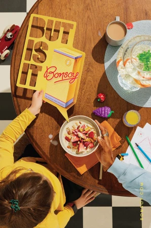 Overhead view of child and adult hands on a wooden table with cereal bowl, Bonsoy carton speech bubble, drinks, and drawing materials.