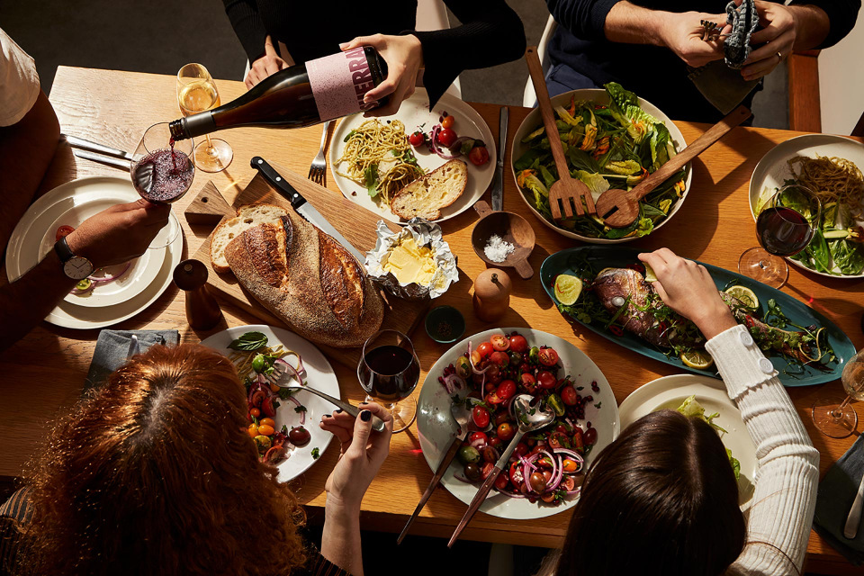 Overhead view of a dining table set with plates of food, wine glasses, cutlery, and a bottle of red wine being poured during a meal.