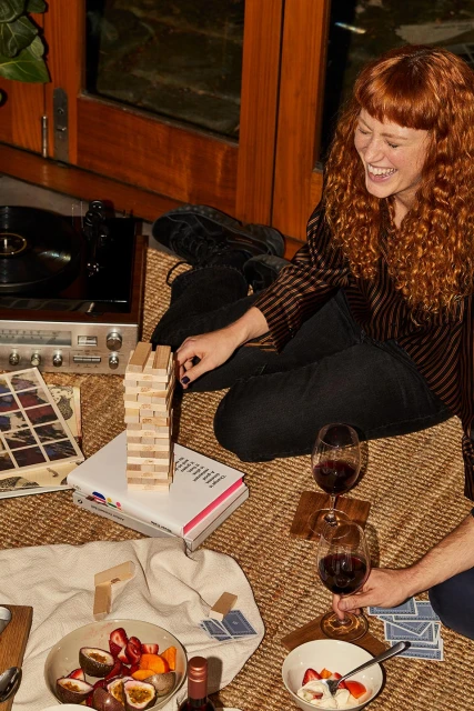 Woman laughing while playing Jenga with friends. Two wine glasses, fruit, vinyl player, and records add to the indoor picnic.