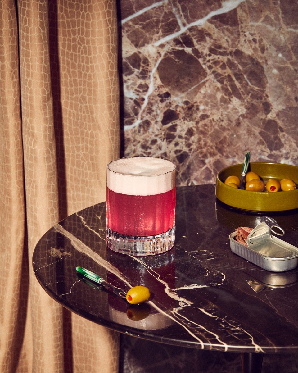 A pink cocktail with white foam in a clear glass on a black marble table, accompanied by olives and a tin of fish.