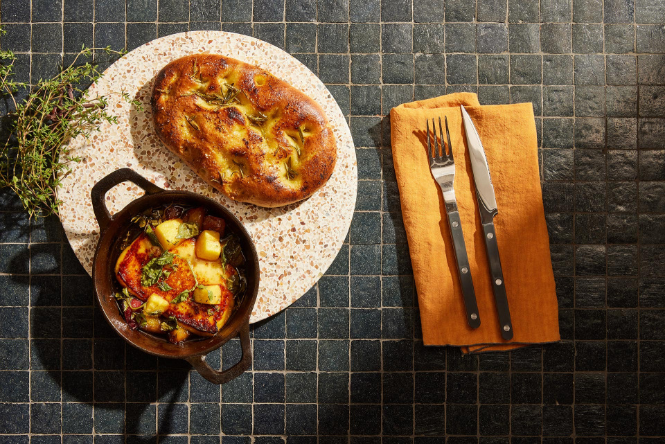 Overhead shot of focaccia bread and potatoes in a cast iron skillet, arranged on a round terrazzo tray, accompanied by a knife and fork.