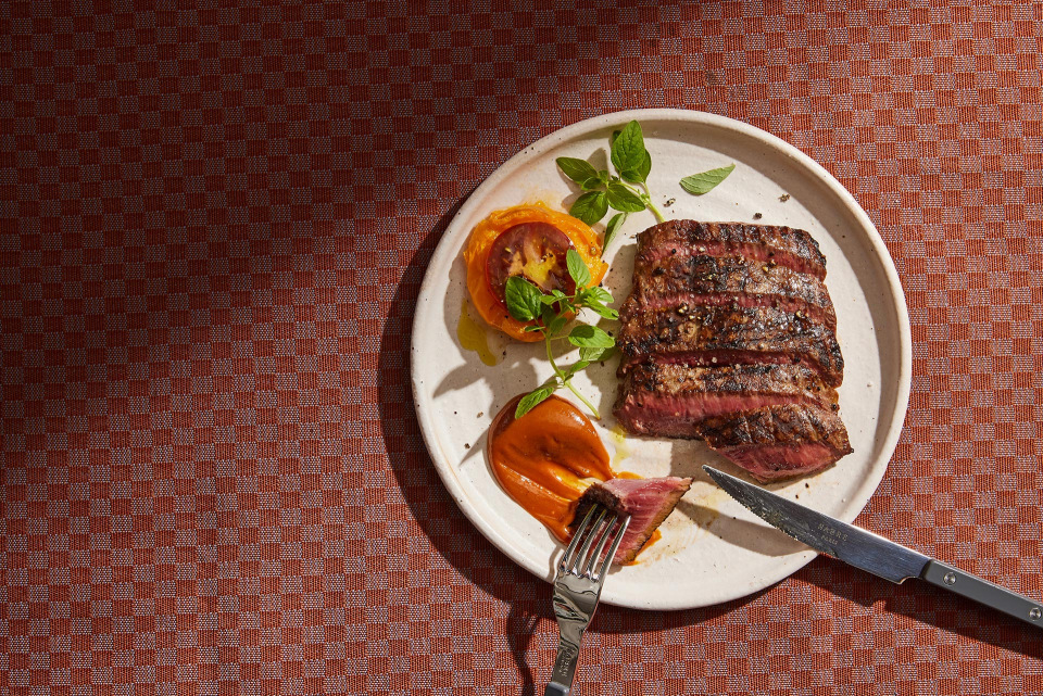 Overhead shot of sliced grilled steak and tomato appetizer on a white plate with a fork and knife, sitting on a patterned tablecloth.