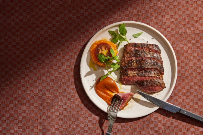 Overhead shot of sliced grilled steak and tomato appetizer on a white plate with a fork and knife, sitting on a patterned tablecloth.
