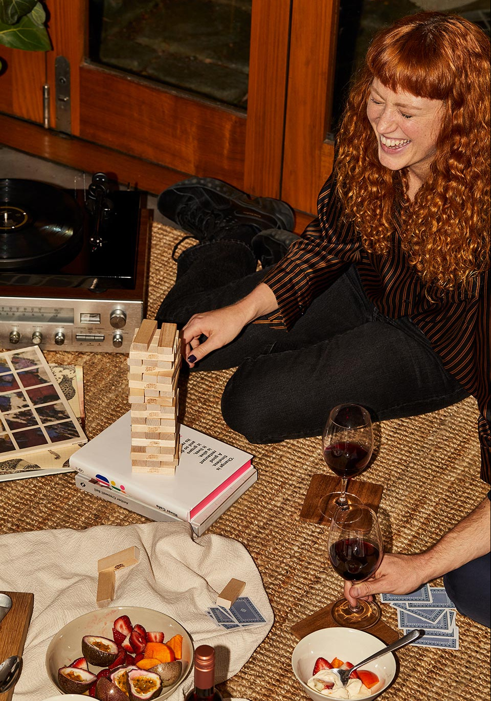 Smiling red-haired woman plays Jenga indoors with fruit, wine, and a record player present in a cozy, warm-toned setting.