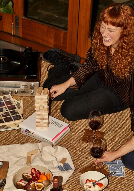 Smiling red-haired woman plays Jenga indoors with fruit, wine, and a record player present in a cozy, warm-toned setting.
