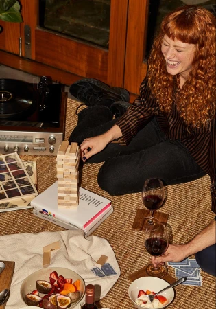 Smiling red-haired woman plays Jenga indoors with fruit, wine, and a record player present in a cozy, warm-toned setting.