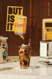 Iced coffee in a tall glass with swirling milk, a hanging yellow Bonsoy sign, jelly beans in a jar, and diner interior props