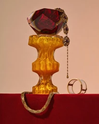 Faceted red glass object atop a textured amber glass pedestal with gold jewelry on a red surface