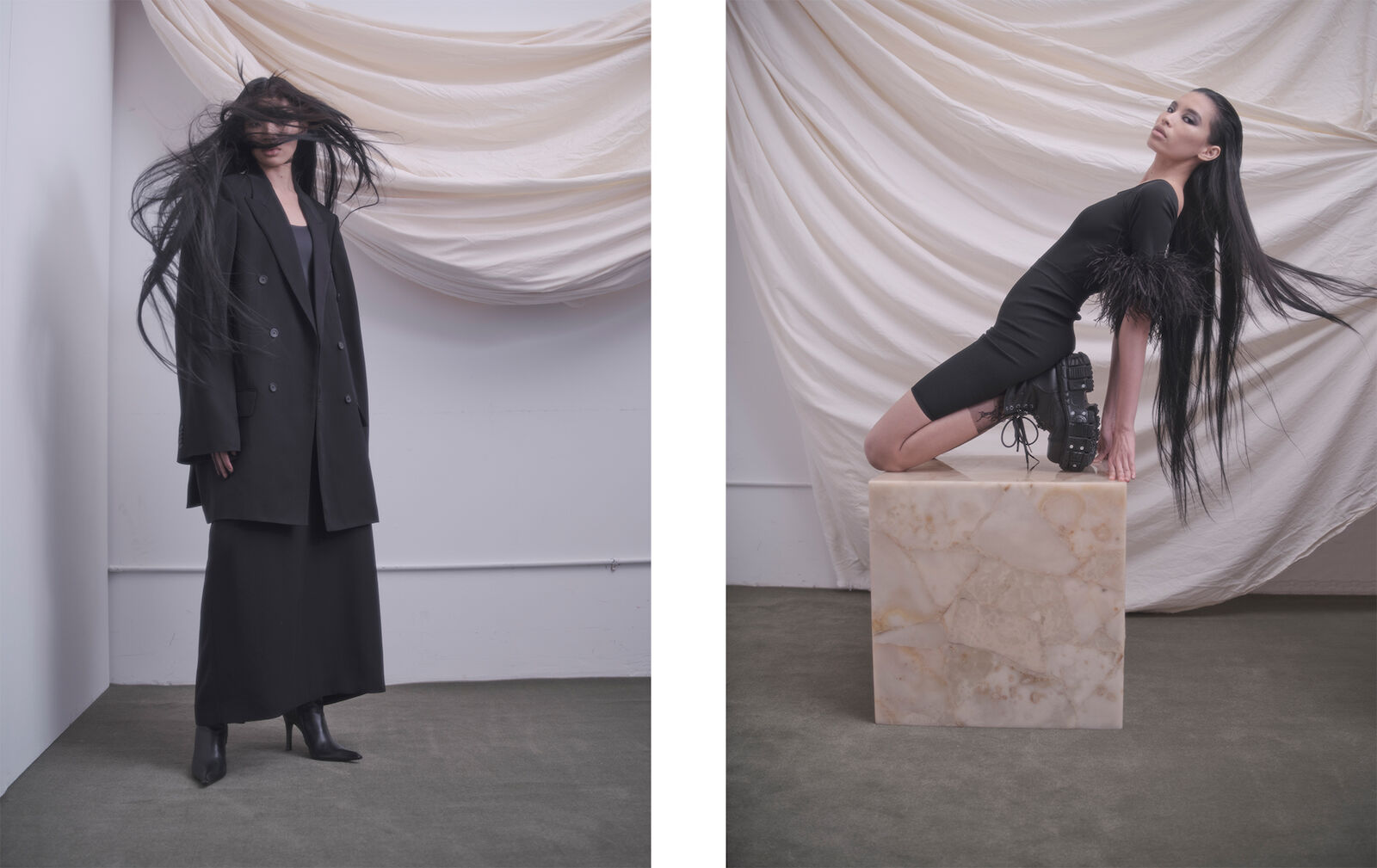 Two women in black outfits posing in front of a draped backdrop. One wears an oversized blazer and skirt, while the other kneels on a marble cube.