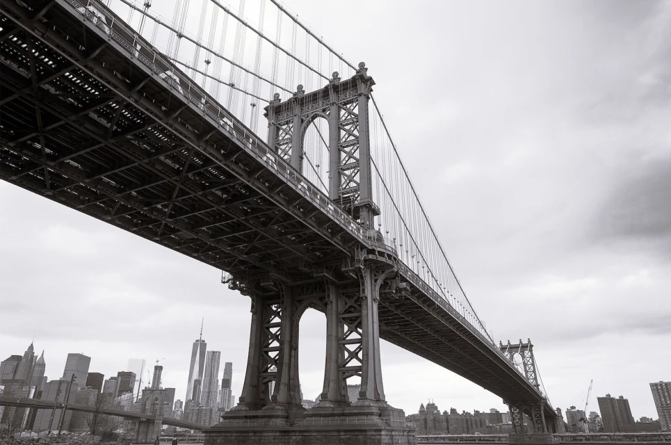 Monochrome view from below of the Manhattan Bridge with the New York City skyline featuring skyscrapers and a cloudy sky in the background.