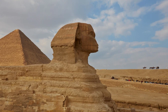 The Great Sphinx of Giza in Egypt, standing near a pyramid, with tourists visible in the background on a sunny day with cloudy skies.