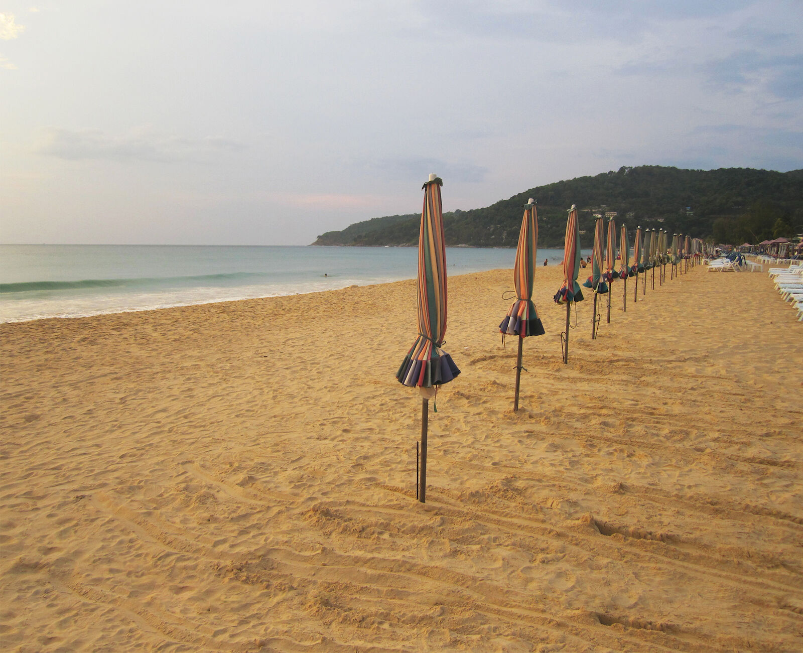 Beach scene featuring numerous closed beach umbrellas in a row along a sandy shore, with the ocean and distant hills in the background.