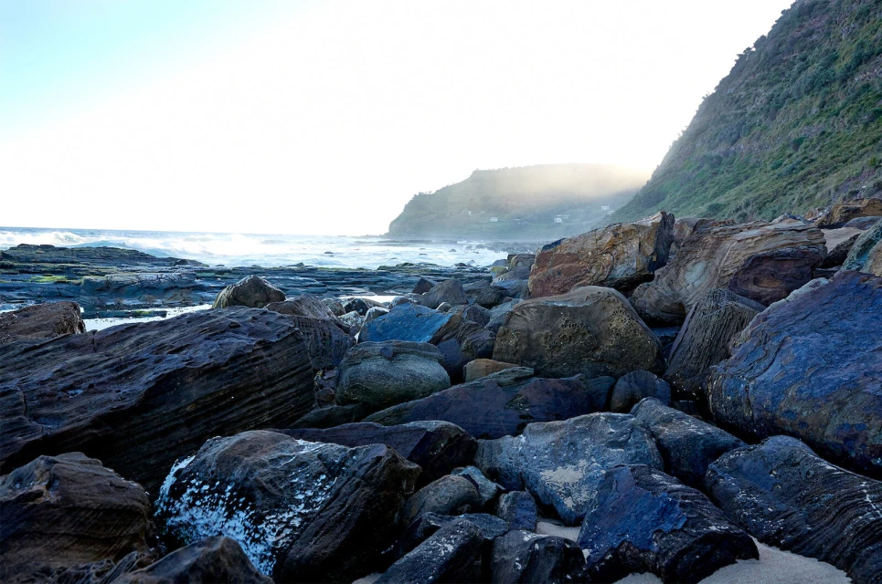 Seascape featuring rugged rocks and waves meeting the coastline on a sunny day, with a lush green hillside in the background.