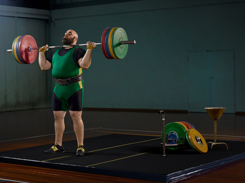 A weightlifter with a beard lifts a barbell with weights in a gym, wearing a green and yellow suit with a weightlifting belt.