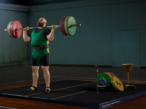 A weightlifter with a beard lifts a barbell with weights in a gym, wearing a green and yellow suit with a weightlifting belt.