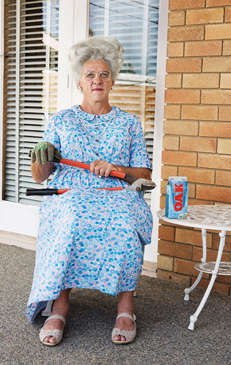 Elderly woman in a floral dress seated on a porch holding pipe wrenches and wearing gloves, with a drink can on a table.