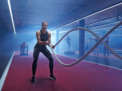 Athletic woman works out with battle ropes in fitness center surrounded by other people exercising in the smoky, neon-lit gym.