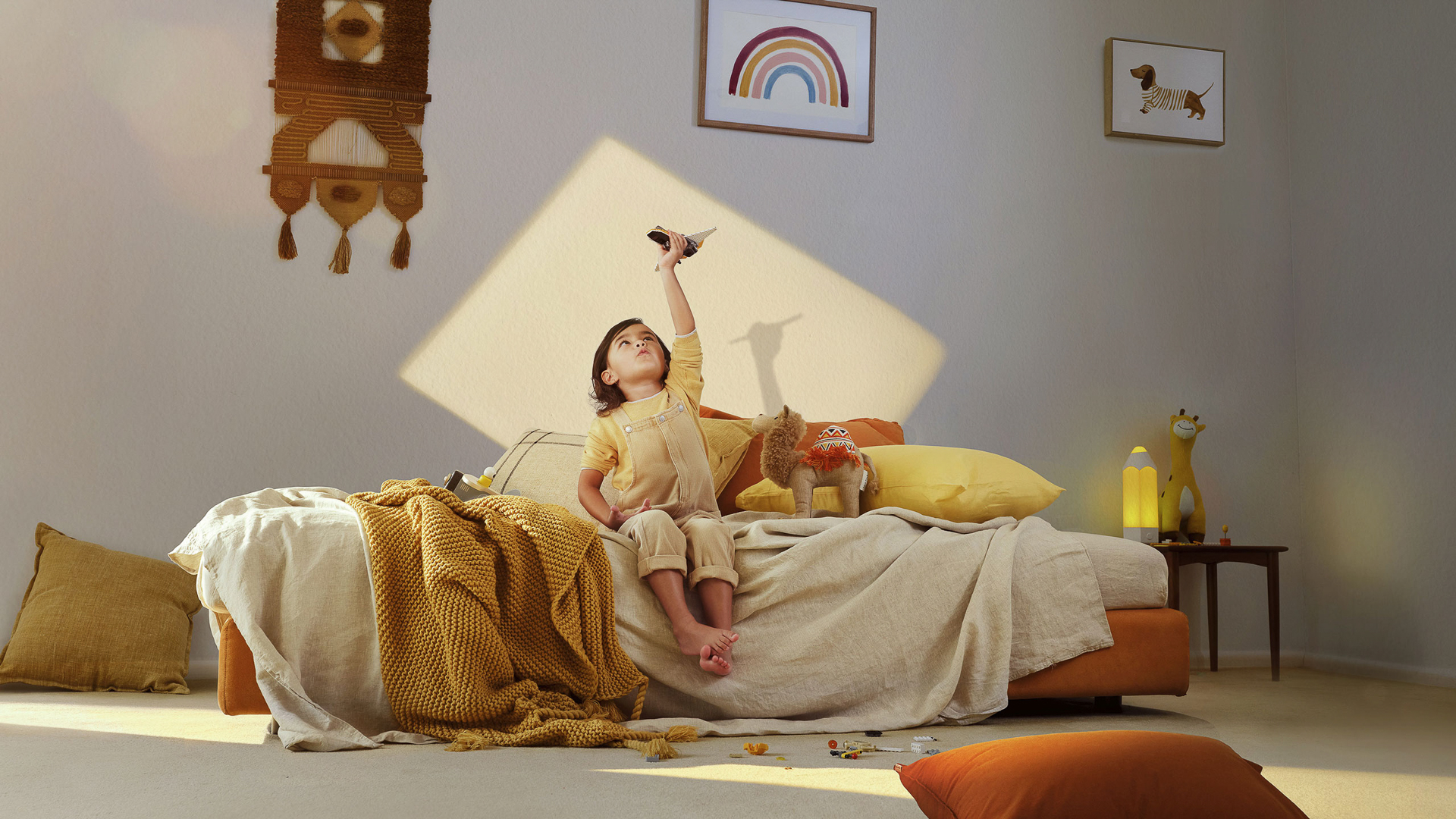 Child playing with toy airplane on a bed with decorative pillows and blankets, bathed in soft sunlight, in a cozy bedroom setting.