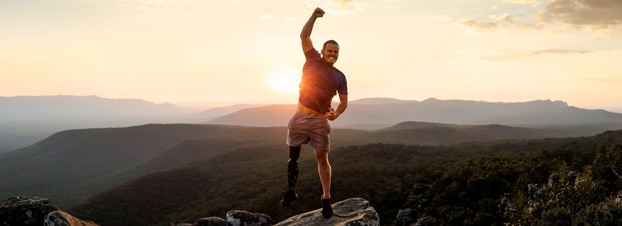 Man with prosthetic leg celebrates on rocky mountaintop at sunset, silhouetted against sky and hills. Sport, disability, accomplishment.