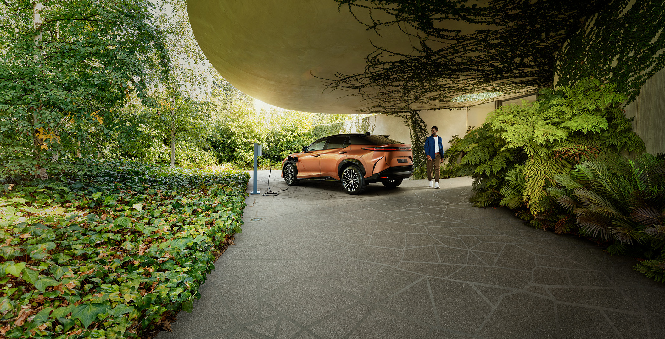 Copper-colored electric SUV parked outdoors near a charging station, with a Black adult male standing to the side, surrounded by greenery.