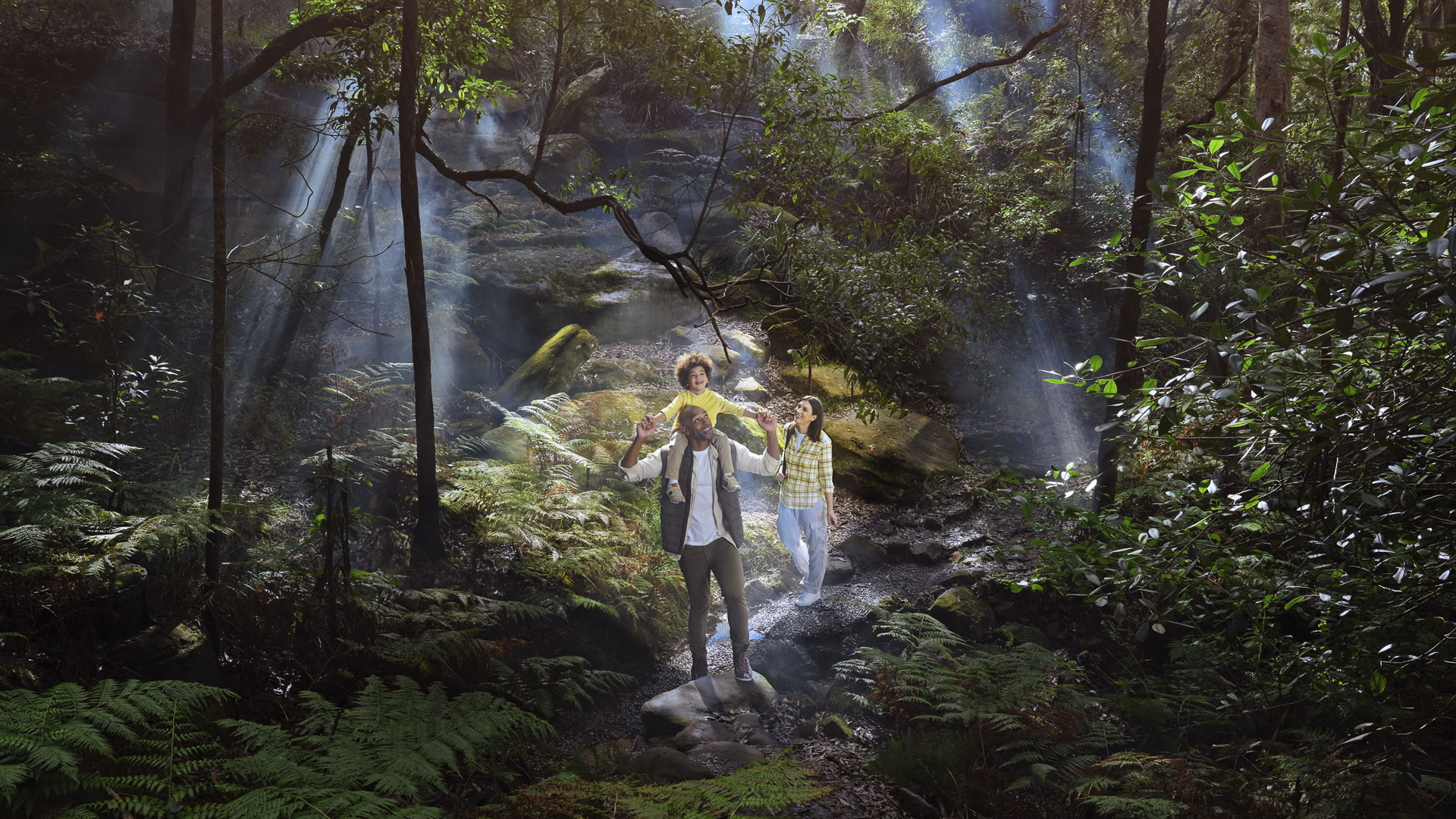 Family of three hiking through sunlit forest. Father with child on shoulders leads the way, followed by mother on a rocky path.
