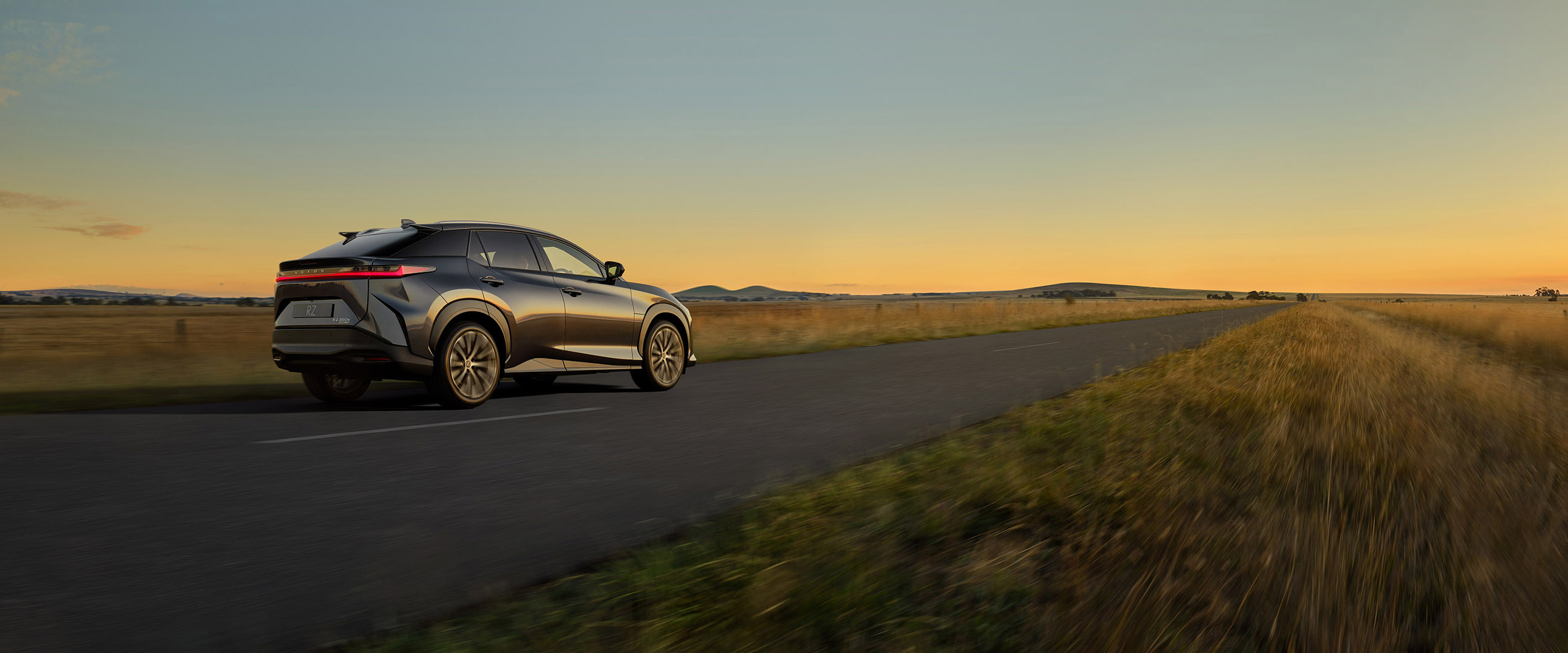 A modern gray SUV drives on a gray asphalt road, with a rural landscape and golden sky in the background at sunset.