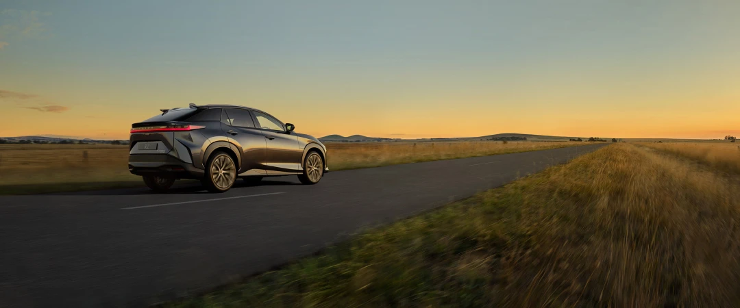 A modern gray SUV drives on a gray asphalt road, with a rural landscape and golden sky in the background at sunset.