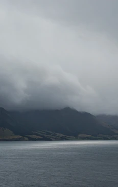 Moody landscape featuring a dark lake reflecting overcast sky, silhouetted mountains in the distance, and diffused light creating a tranquil scene.