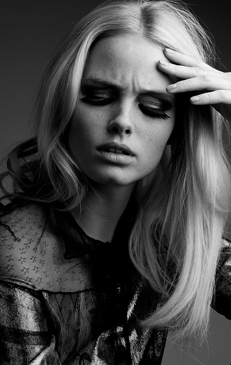 Black and white studio portrait of a Caucasian female model with heavy eye makeup and blond hair, wearing a sheer, patterned top.