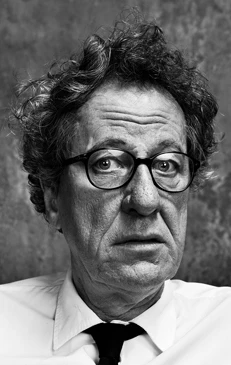 Portrait of older man wearing glasses, white collared shirt, and dark tie against textured grey background.