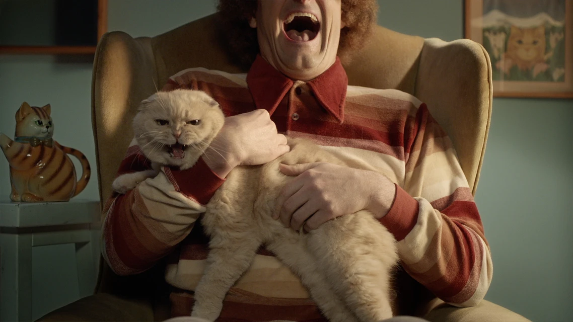 A man with curly red hair sits in a chair while holding a grumpy-looking cat with its mouth open. A cat teapot sits on a nearby table.