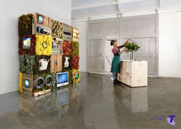 Woman arranging flowers at a light wooden counter beside a large modular wall installation of plants and screens inside a warehouse.