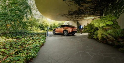 Copper electric crossover car parked under a cantilevered concrete roof surrounded by lush ferns and a man standing nearby.
