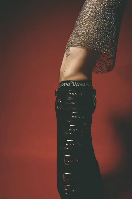 Cropped studio shot of an adult wearing black leather pants with buckles, a silver metallic top, and underwear; set against a red backdrop.