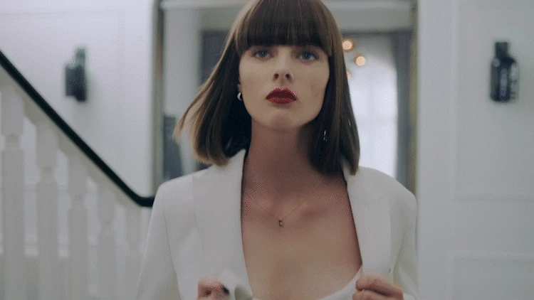 Elegant adult woman in white blazer and dark lipstick walking in brightly lit hallway with neutral decor and dark stair railing.