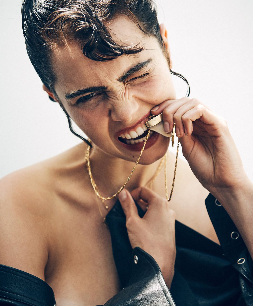 Close-up portrait of a woman with dark hair biting a necklace, wearing a leather jacket and a bold expression against a white backdrop.