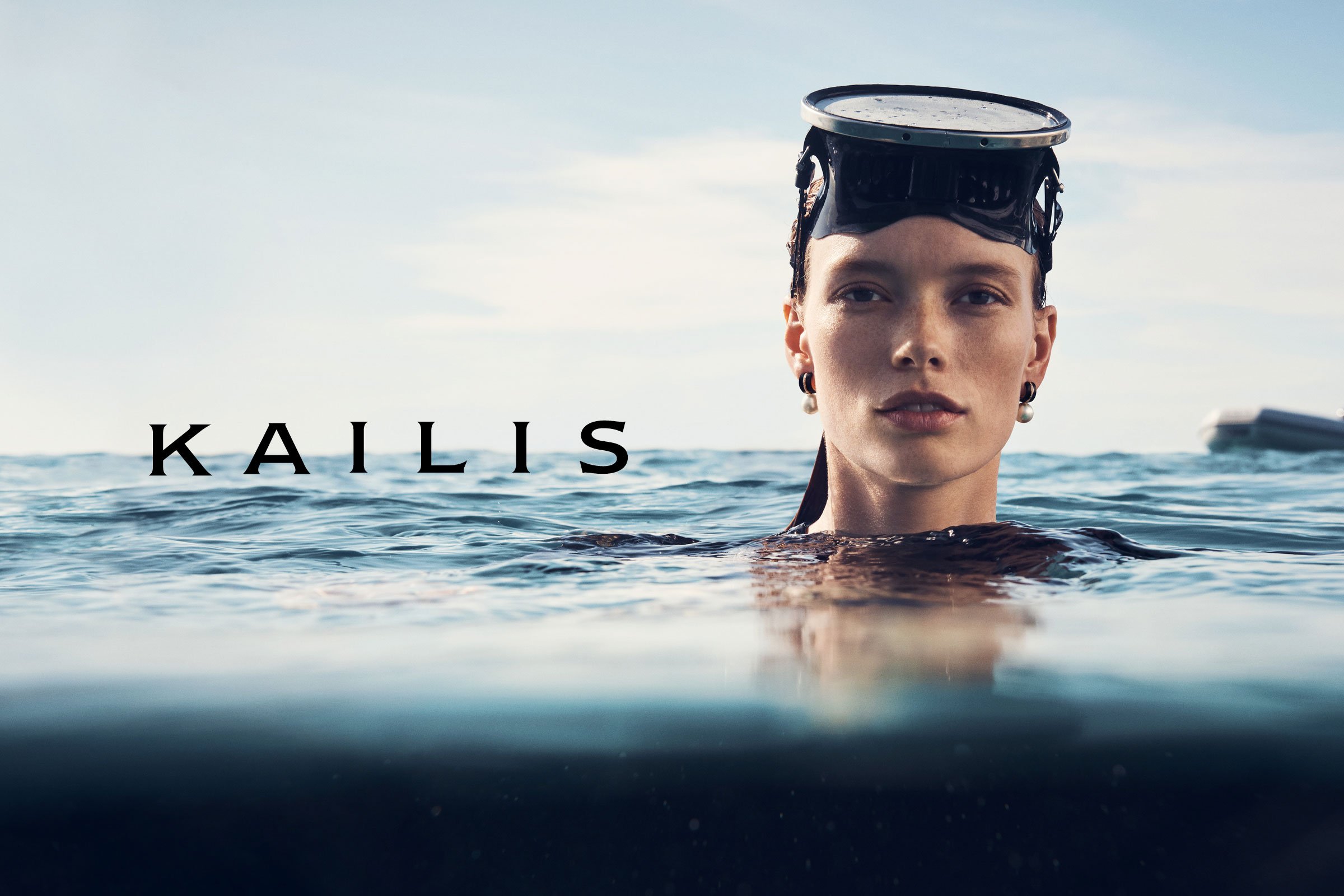 Close-up of a woman in the water wearing a diving mask and pearl earrings; 'KAILIS' text overlayed on the image against the ocean backdrop.