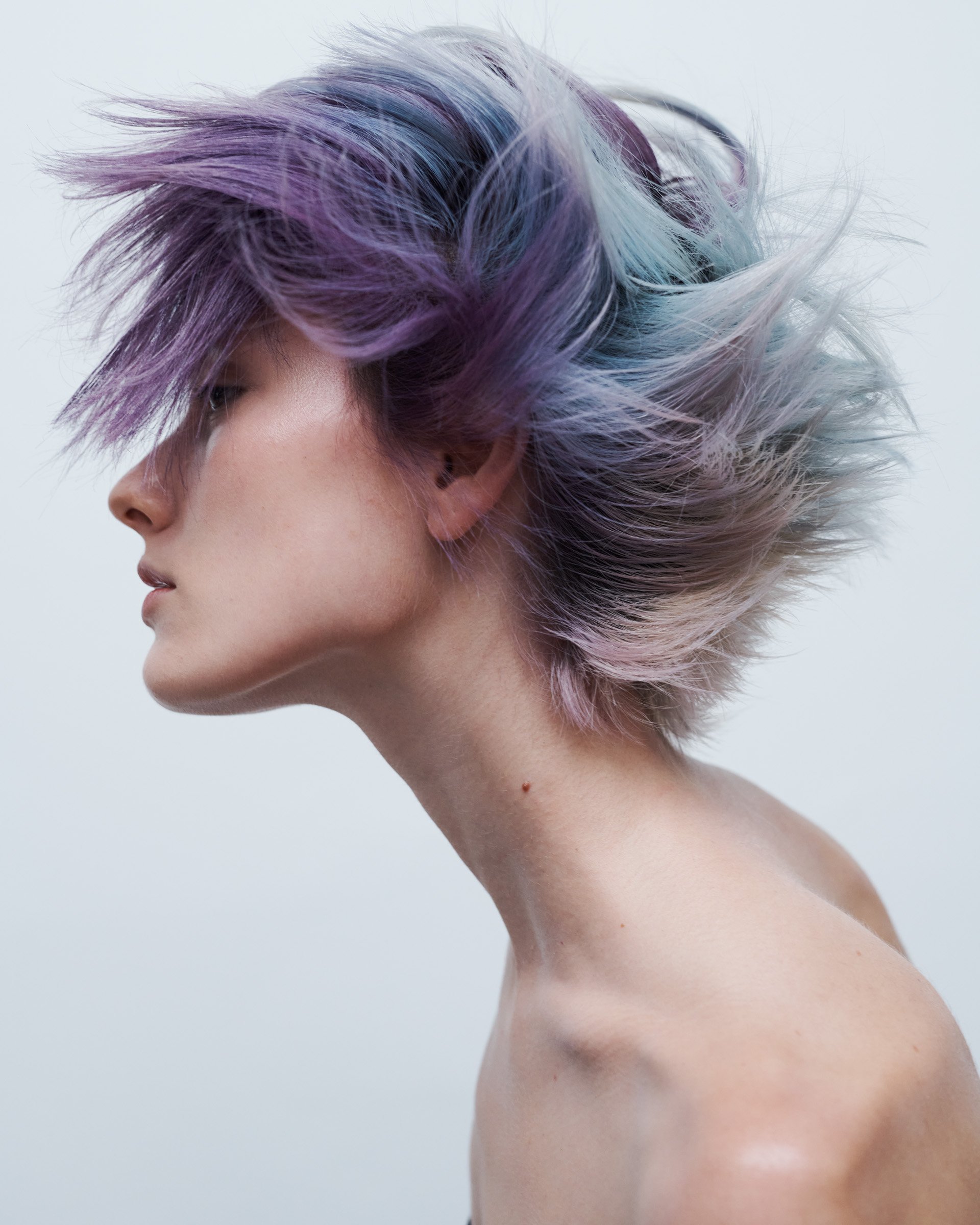 Profile shot of a stylish woman with trendy, choppy layered short hair dyed in pastel purple, blue, and pink tones against a light backdrop.